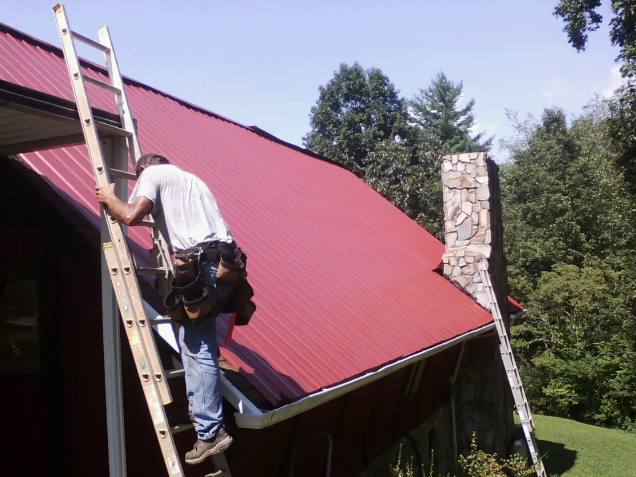 Roofer on a ladder beside a red metal roof and stone chimney — Soffit & Fascia Repair by Roofing Brothers in West Monroe