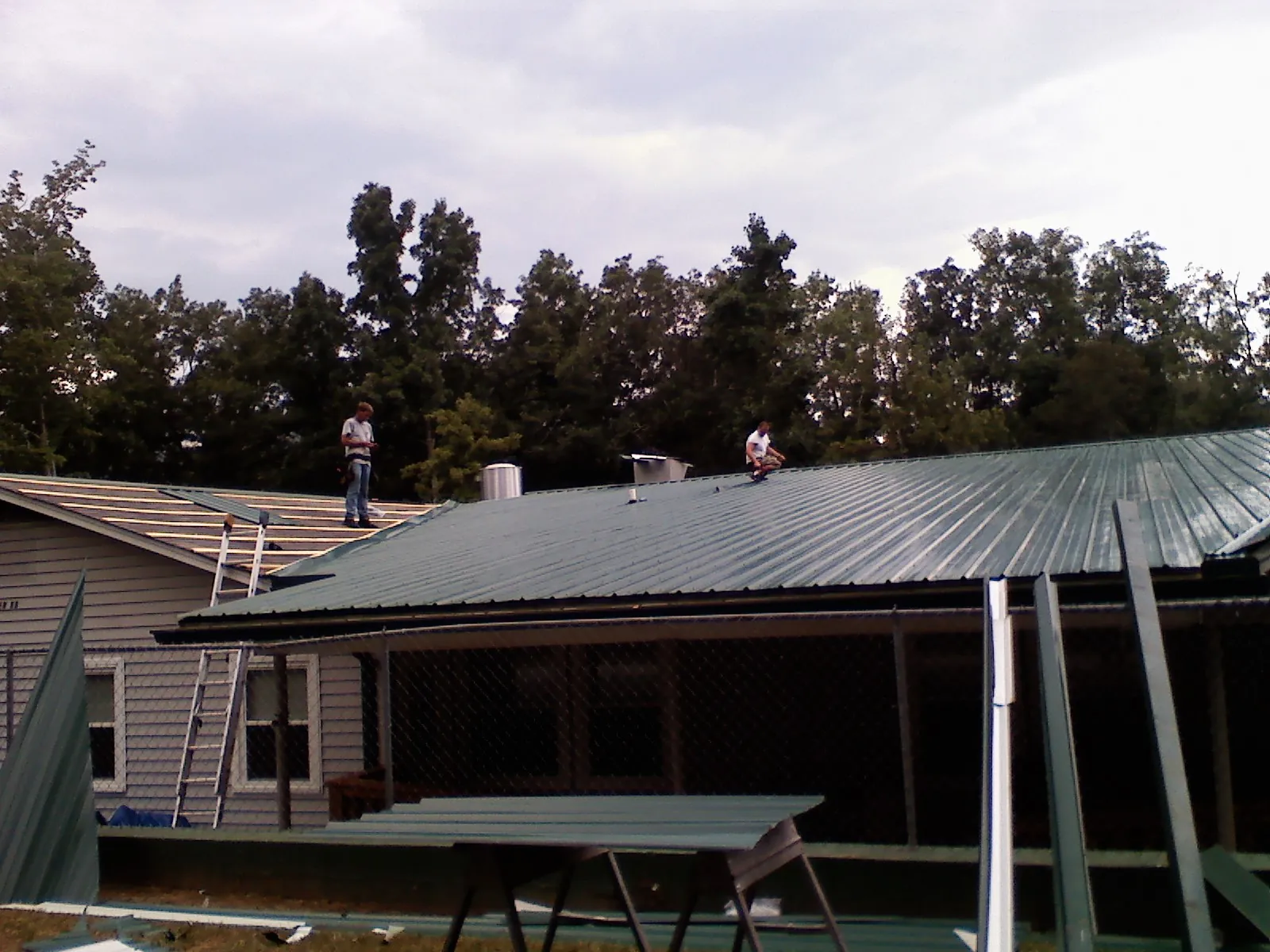 Two roofers installing green metal panels above a covered porch in West Monroe